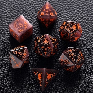 May include: Set of seven black and orange dice with Halloween-themed designs. The dice are made of a translucent material with a glittery finish. The numbers on the dice are in orange and the designs are in black.
