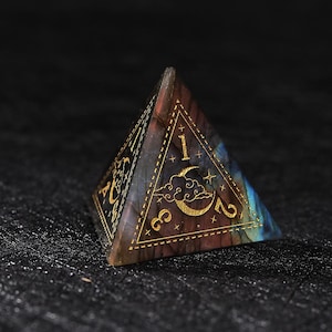 May include: A pyramid-shaped, multi-colored gemstone die with gold-colored numbers and celestial designs. The die features the numbers 1, 2, and 3, along with a crescent moon and stars. The gemstone has a mix of blue, brown, and purple hues.