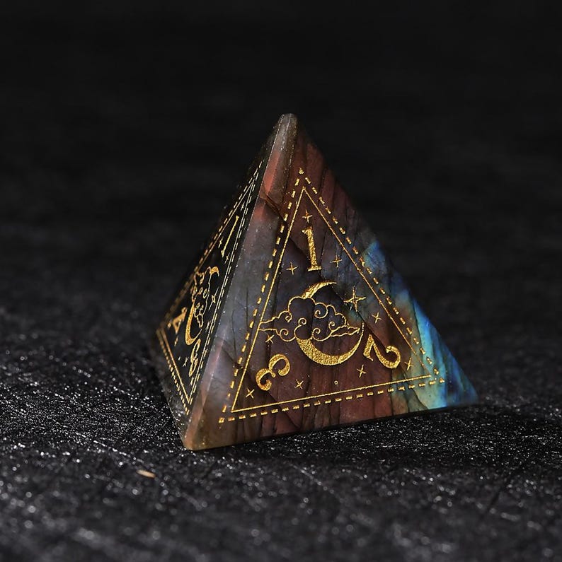 May include: A pyramid-shaped, multi-colored gemstone die with gold-colored numbers and celestial designs. The die features the numbers 1, 2, and 3, along with a crescent moon and stars. The gemstone has a mix of blue, brown, and purple hues.