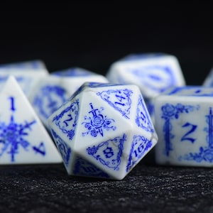 May include: A set of polyhedral dice featuring a white base with intricate blue floral and sword patterns. The twenty-sided die in the foreground displays a central rose and sword emblem, with numbers printed in a dark blue gothic-style font.