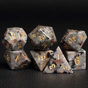 Peut inclure: Ensemble de sept d&eacute;s mouchet&eacute;s noirs et gris avec des chiffres dor&eacute;s. Les d&eacute;s sont de formes vari&eacute;es, notamment un cube, une pyramide et un dod&eacute;ca&egrave;dre.