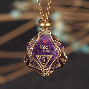 May include: A gold necklace with a purple amethyst gemstone d20 dice pendant. The dice has gold accents and the inscription "K&L 11.11.2025" and a small red heart.