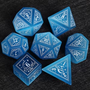 Puede incluir: Un conjunto de siete dados poliédricos azules con numeración blanca y diseños decorativos. Los dados tienen varias formas geométricas, incluyendo un d4, d6, d8, d10, d12, d20 y un dado de porcentaje. Los dados están dispuestos sobre una superficie oscura.