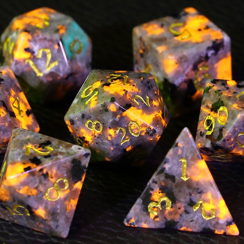 Peut inclure: Ensemble de sept d&eacute;s poly&eacute;driques en pierre mouchet&eacute;e de gris fonc&eacute; et de blanc avec des chiffres dor&eacute;s. Les d&eacute;s brillent dans le noir d'une couleur orange vif.
