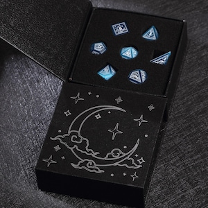 May include: A set of seven blue and white polyhedral dice in a black box. The dice are displayed in a black foam insert. The box has a black lid with a crescent moon and star design.