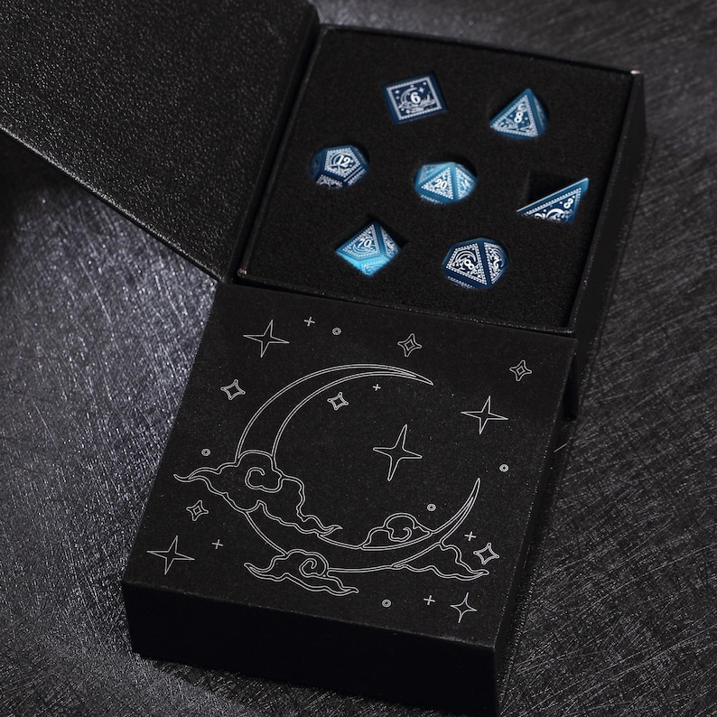 May include: A set of seven blue and white polyhedral dice in a black box. The dice are displayed in a black foam insert. The box has a black lid with a crescent moon and star design.