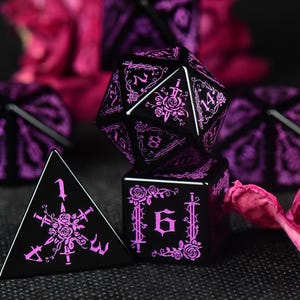 Pode incluir: Um conjunto de dados poliédricos pretos com gravações cor-de-rosa complexas de rosas e espadas. O conjunto inclui um dado de vinte lados, um de quatro e um de seis, todos exibindo números claros e motivos florais e de lâminas detalhados.