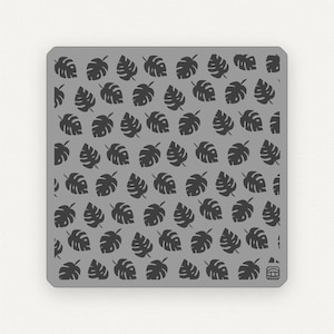 Puede incluir: Una plantilla de metal gris con un patrón repetitivo de hojas tropicales negras. La plantilla es cuadrada y tiene un pequeño logotipo en la esquina inferior derecha.