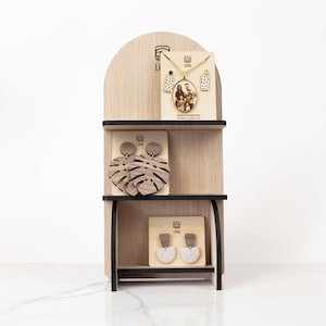 Puede incluir: Expositor de joyería de madera con tres estantes. El estante superior presenta un collar de oro con un colgante de foto y aretes a juego. El estante del medio tiene un par de aretes de madera con forma de hojas de monstera. El estante inferior tiene un par de aretes de arcilla blanca.