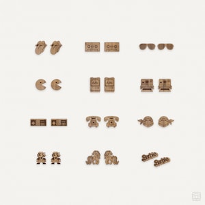 Könnte beinhalten: Ein Set mit 10 Paar Holzohrringen mit verschiedenen Popkultur-Ikonen, darunter ein Kassettenband, eine Zunge, ein Pac-Man, eine Polaroidkamera, ein Telefon, eine Teenage Mutant Ninja Turtle, ein Super Mario-Charakter, ein My Little Pony und das Wort "Barbie".