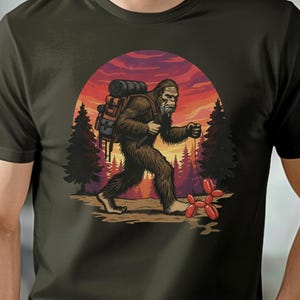 以下が含まれることがあります： ビッグフットのグラフィックが特徴のダークオリーブグリーンのTシャツ。イラストは、バックパックを背負ってハイキングするビッグフット、夕日の背景、赤い風船の動物を描いています。このシャツは柔らかい素材で作られています。