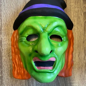 May include: A green witch mask with orange hair, a black hat with a purple band, and open eye and mouth holes. The mask has a wrinkled face and is made of a smooth material. The mask is for Halloween.