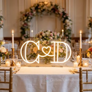 Pode incluir: Uma placa de néon com as letras "C & D" e um símbolo de coração no centro, iluminada por uma luz branca quente. A placa está colocada numa mesa de casamento, com velas e arranjos florais. O texto "Custom Neon Wonderland" está na parte inferior.