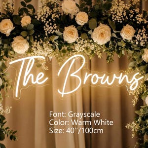 Puede incluir: Un letrero de ne&oacute;n blanco c&aacute;lido que muestra el texto The Browns en una fuente cursiva, montado dentro de un arco floral de rosas crema y follaje verde. El letrero presenta la fuente Grayscale, color blanco c&aacute;lido y mide 100 cm de ancho.