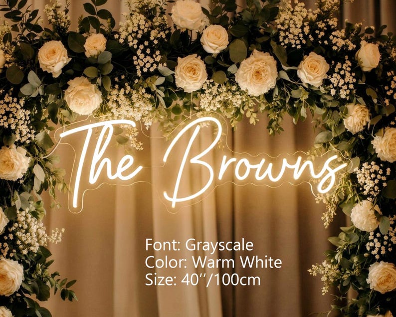 Puede incluir: Un letrero de ne&oacute;n blanco c&aacute;lido que muestra el texto The Browns en una fuente cursiva, montado dentro de un arco floral de rosas crema y follaje verde. El letrero presenta la fuente Grayscale, color blanco c&aacute;lido y mide 100 cm de ancho.