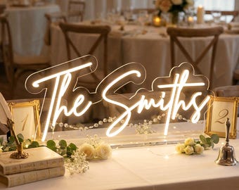 Puede incluir: Un letrero de neón personalizado con el texto The Smiths en una fuente cursiva blanca brillante, montado sobre un soporte de acrílico transparente. Se exhibe en una mesa con flores blancas y follaje verde decorativo.