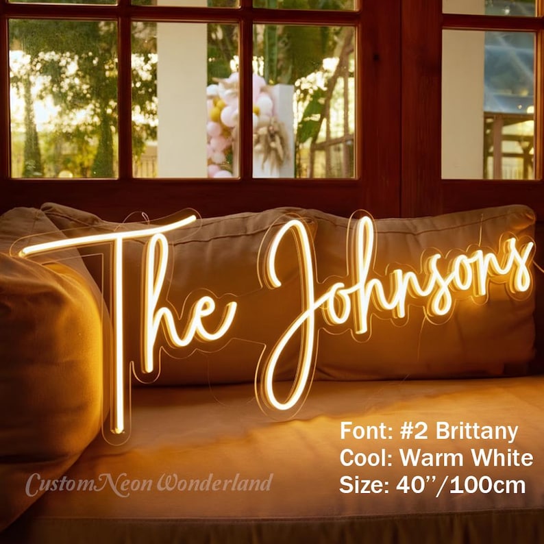 Puede incluir: Un letrero de ne&oacute;n blanco c&aacute;lido en letra cursiva que dice The Johnsons. El letrero mide 100 cm de ancho y utiliza la fuente Brittany. Se muestra sobre un coj&iacute;n beige, con detalles de texto visibles en la esquina inferior derecha.