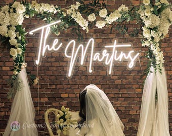 Puede incluir: Un letrero de neón blanco brillante que muestra el texto The Martins en una fuente cursiva. El letrero está montado contra una pared de ladrillo marrón rústico y está enmarcado por un arco decorado con flores blancas, follaje verde y tela blanca.