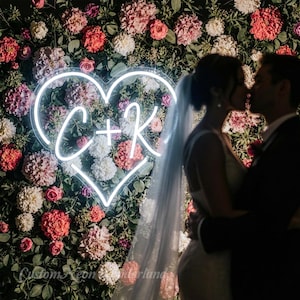 May include: A neon heart with the letters "C + K" against a backdrop of flowers in shades of pink, red, and white. A couple is kissing in front of the floral arrangement.