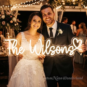 Puede incluir: Un letrero de ne&oacute;n personalizado con el texto The Wilsons en una fuente cursiva de color blanco c&aacute;lido brillante, que termina con un contorno en forma de coraz&oacute;n. El letrero est&aacute; hecho de una base de acr&iacute;lico transparente y emite luz brillante.