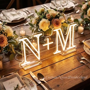 May include: A wooden table is set for a special occasion, featuring floral arrangements, candles, and place settings. The centerpiece is a neon sign displaying the letters "N + M" in a warm, inviting glow. The sign is from "Custom Neon Wonderland."