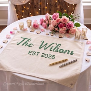May include: A rectangular cream-colored fabric banner featuring the dark green embroidered text "The Wilsons" and "EST 2026". The banner includes metal grommets in the top corners and is displayed with a gold pen and a silver pen resting on its surface.