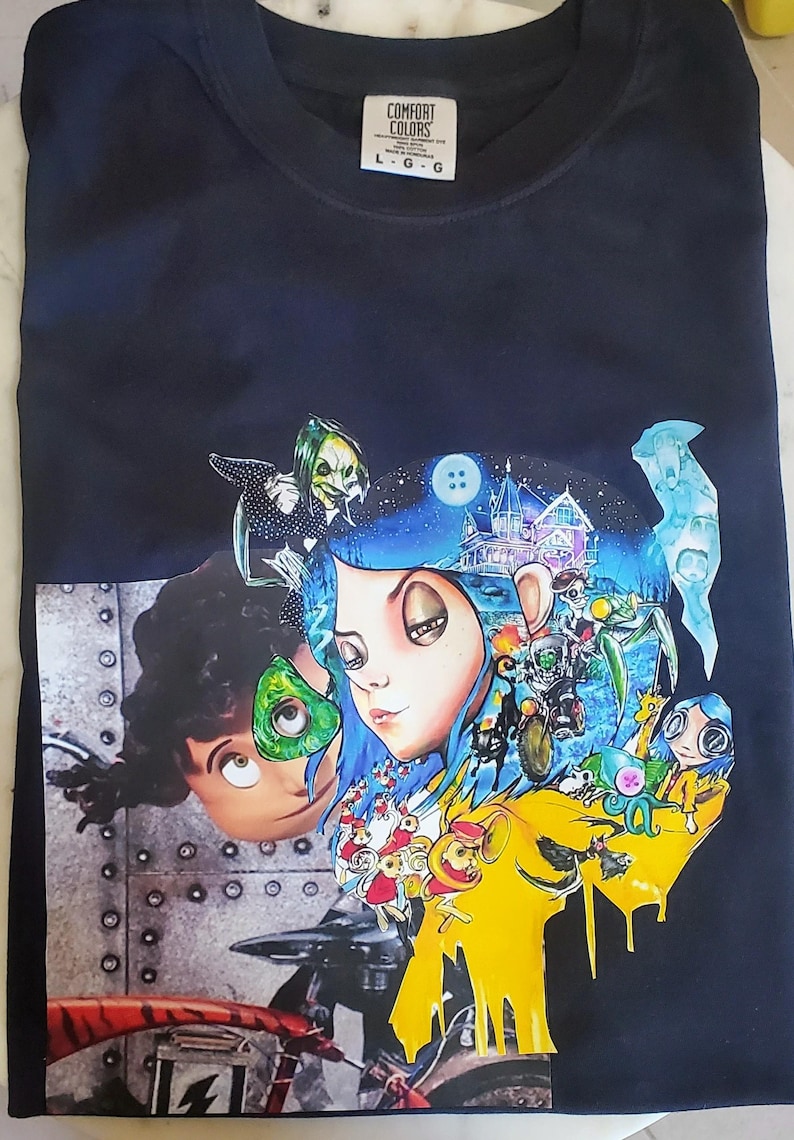 May include: Black Comfort Colors t-shirt featuring a colorful graphic print. The print includes cartoon characters with blue, green, and yellow accents. The shirt has a crew neck and the Comfort Colors logo.