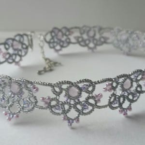 May include: A silver-colored choker necklace with a delicate, openwork design. The necklace features small, light pink and lavender-colored beads. The intricate pattern and beadwork create an elegant and feminine accessory.