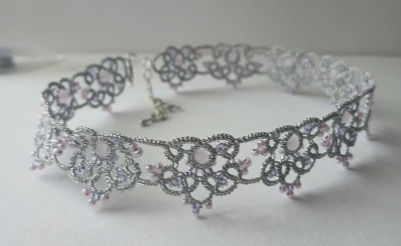 May include: A silver-colored choker necklace with a delicate, openwork design. The necklace features small, light pink and lavender-colored beads. The intricate pattern and beadwork create an elegant and feminine accessory.