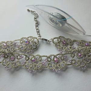 May include: Silver-toned choker necklace with intricate lace-like detailing. The necklace features iridescent pink gemstones and small, light purple beads. A silver chain and clasp are visible, along with a clear tag.