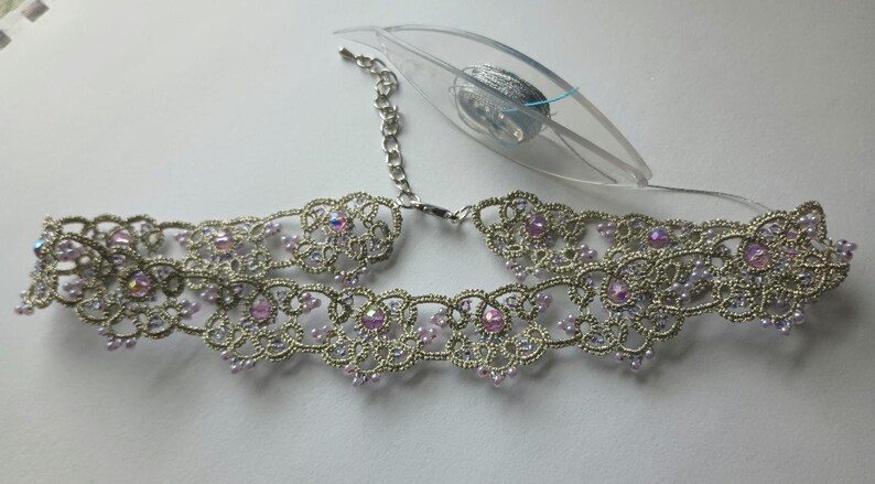 May include: Silver-toned choker necklace with intricate lace-like detailing. The necklace features iridescent pink gemstones and small, light purple beads. A silver chain and clasp are visible, along with a clear tag.