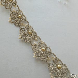 May include: A delicate, silver-toned lace bracelet with a floral design. The bracelet features small, round, pearl-like beads and larger, golden-colored beads at the center of each floral motif. The intricate lace pattern is visible.