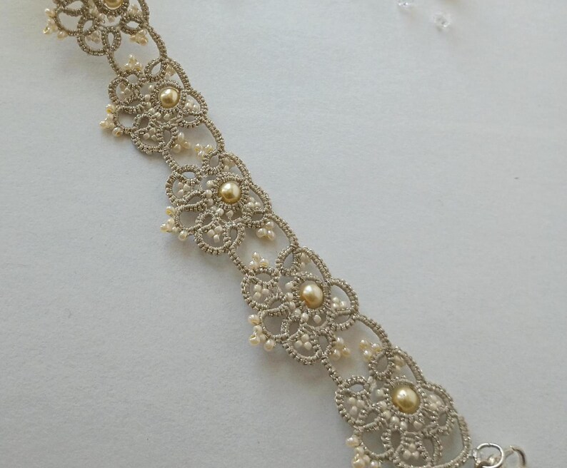 May include: A delicate, silver-toned lace bracelet with a floral design. The bracelet features small, round, pearl-like beads and larger, golden-colored beads at the center of each floral motif. The intricate lace pattern is visible.