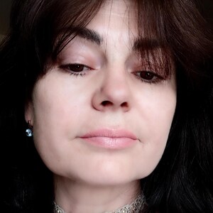 May include: A close-up portrait of a person wearing a silver lace choker necklace with pearl accents and a small blue gemstone earring. The person has dark hair and is looking down. The skin tone is fair, and the lips are a light pink.