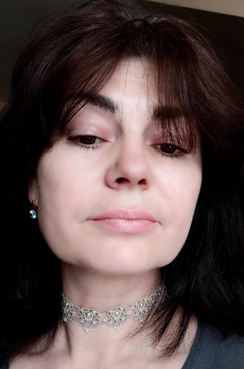 May include: A close-up portrait of a person wearing a silver lace choker necklace with pearl accents and a small blue gemstone earring. The person has dark hair and is looking down. The skin tone is fair, and the lips are a light pink.