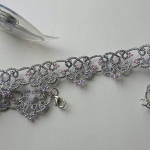 May include: A silver-colored, lace-like choker necklace with small, light pink beads. The necklace has a delicate floral design and a clasp closure. A needle and thread are in the background.