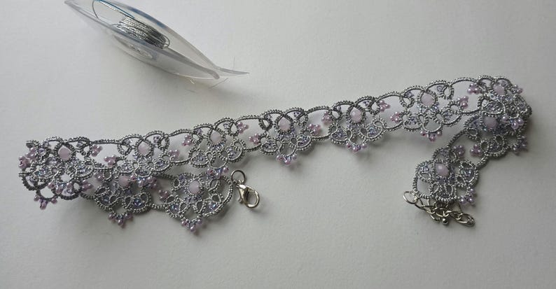 May include: A silver-colored, lace-like choker necklace with small, light pink beads. The necklace has a delicate floral design and a clasp closure. A needle and thread are in the background.