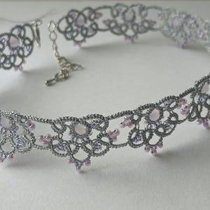 May include: A silver-colored choker necklace with a delicate, lacy design. The necklace features small, light pink and lavender-colored beads. The clasp and chain are also silver. The necklace is designed to be worn around the neck.