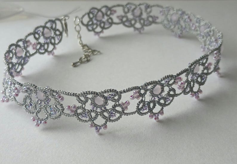 May include: A silver-colored choker necklace with a delicate, lacy design. The necklace features small, light pink and lavender-colored beads. The clasp and chain are also silver. The necklace is designed to be worn around the neck.