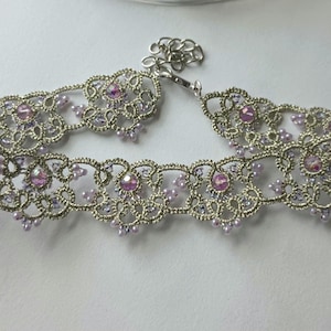 May include: A silver-toned choker necklace with an intricate lace design. The necklace is adorned with small, light purple beads and larger, pink gemstones. The clasp is a simple chain and hook closure. The overall style is delicate and feminine.