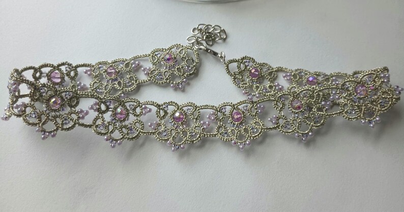 May include: A silver-toned choker necklace with an intricate lace design. The necklace is adorned with small, light purple beads and larger, pink gemstones. The clasp is a simple chain and hook closure. The overall style is delicate and feminine.