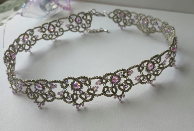 May include: A silver-toned, open-work choker necklace with a delicate floral design. The necklace is adorned with iridescent pink crystals and smaller pink beads, creating a sparkling effect. The clasp is visible at the back.
