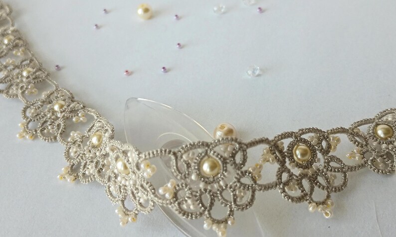 May include: A delicate, silver-colored lace collar with pearl accents. The intricate design features floral patterns and small, round pearls. Scattered pearls and beads are visible in the background, suggesting a bridal or formal theme.