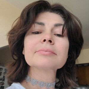 May include: A close-up shot of a person wearing a silver choker necklace. The necklace features a delicate, openwork design with small, light pink accents. The person has dark brown hair and is wearing a white shirt.