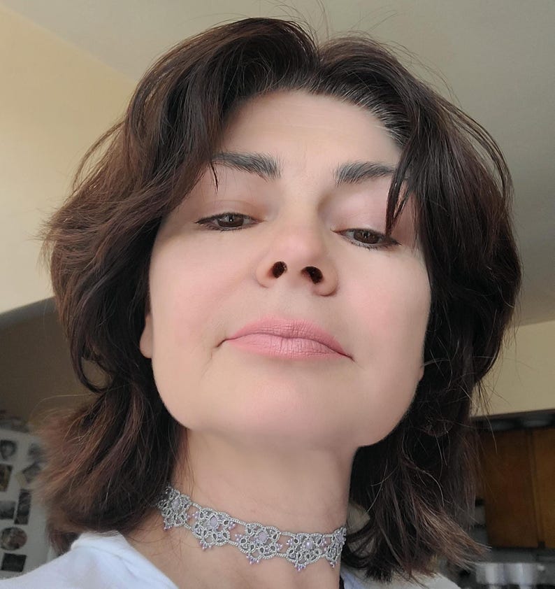 May include: A close-up shot of a person wearing a silver choker necklace. The necklace features a delicate, openwork design with small, light pink accents. The person has dark brown hair and is wearing a white shirt.