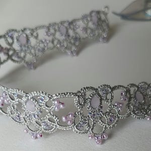 May include: A silver-colored, open-work headband with lavender-colored accents. The delicate design features small, round lavender beads and larger, oval-shaped lavender stones. The headband is likely for special occasions.