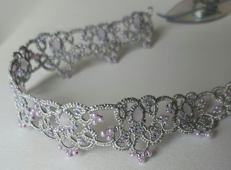 May include: A silver-colored, open-work headband with lavender-colored accents. The delicate design features small, round lavender beads and larger, oval-shaped lavender stones. The headband is likely for special occasions.