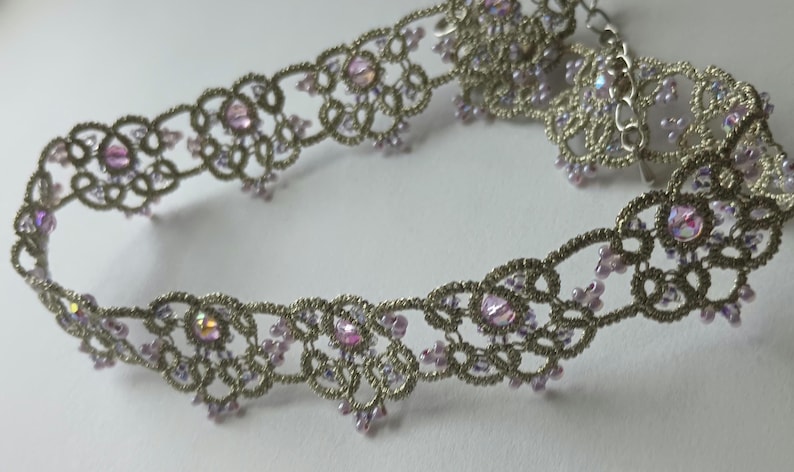 May include: A silver-toned choker necklace with a delicate, openwork design. The necklace is adorned with iridescent pink crystals and smaller pink beads. The intricate lace-like pattern creates a romantic and elegant aesthetic, suitable for special occasions.