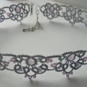 May include: A silver-colored choker necklace with a delicate lace design. The necklace features a repeating pattern of floral shapes and is adorned with small, light purple beads. The clasp and chain are silver-toned.