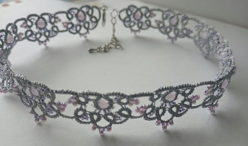 May include: A silver-colored choker necklace with a delicate lace design. The necklace features a repeating pattern of floral shapes and is adorned with small, light purple beads. The clasp and chain are silver-toned.